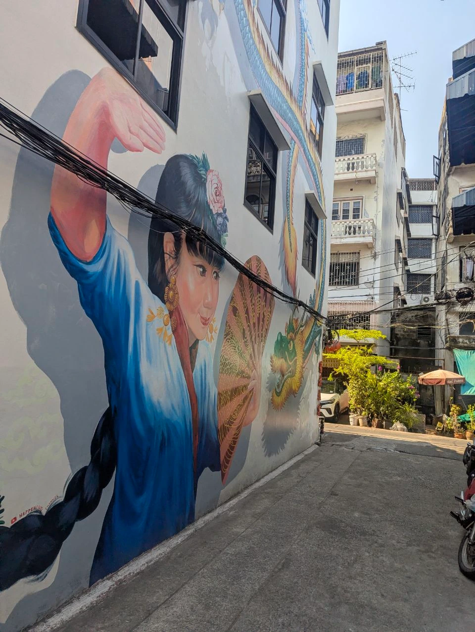 Bangkok Photoshoot Packing - Mural Lane A large mural of a woman holding a fan fills a narrow sunlit Bangkok lane crossed by overhead utility cables.