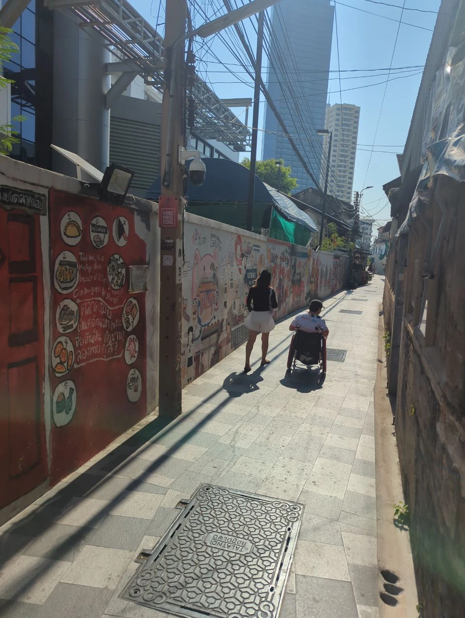 Talat Noi Photoshoot Guide - Mural-Lined Alley Two visitors, one walking and one using a wheelchair, move through a mural-lined Talat Noi alley in Bangkok.
