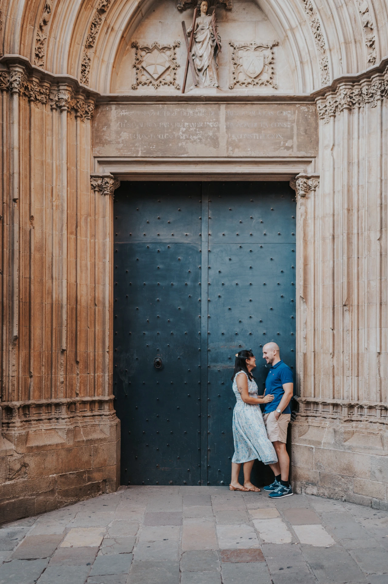The couple embracing beside a blue door beneath an ornate carved stone arch in the Gothic Quarter during their anniversary photoshoot in Barcelona.