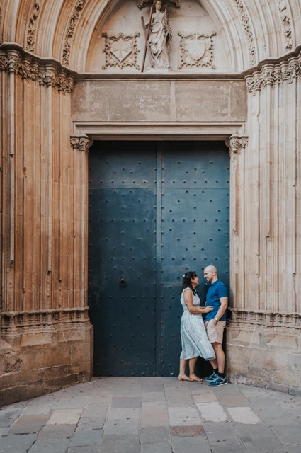 The couple embracing beside a blue door beneath an ornate carved stone arch in the Gothic Quarter during their anniversary photoshoot in Barcelona.