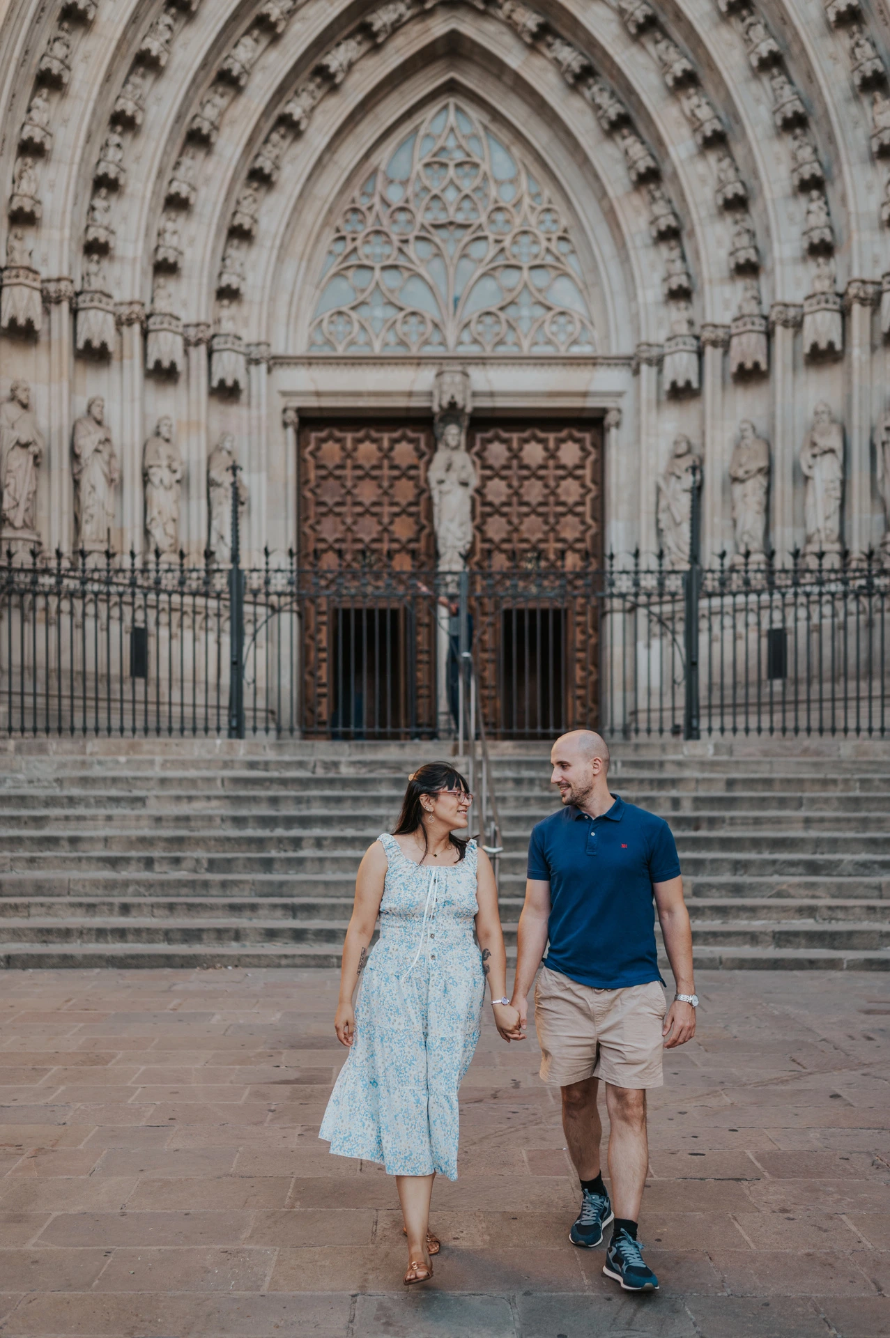 The couple smiling at each other as they walk hand in hand in front of Barcelona Cathedral during their anniversary photoshoot in Barcelona.