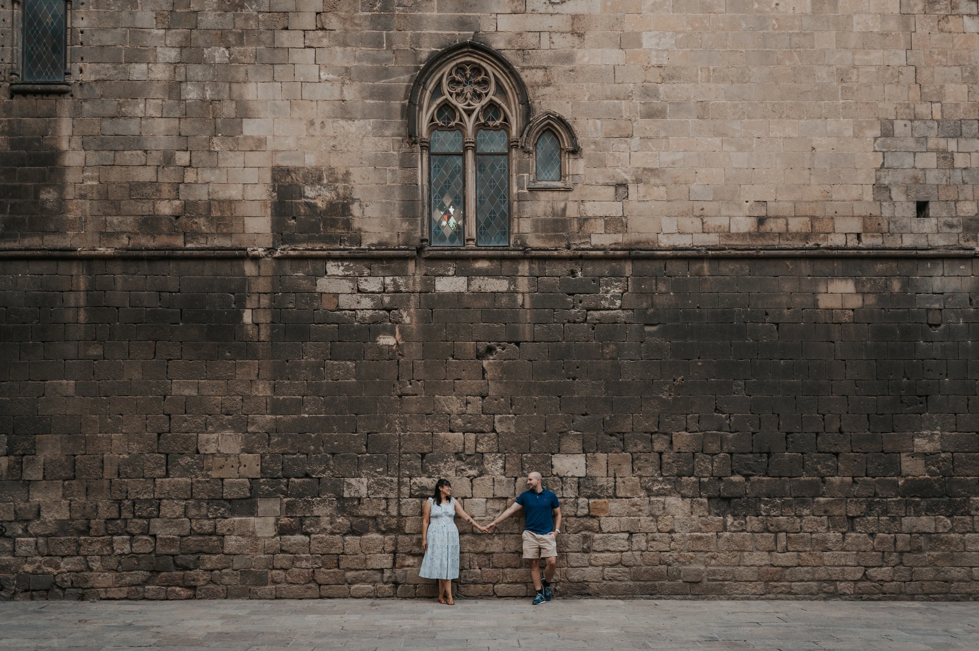 A wide view of the couple standing below a tall historic stone wall with a Gothic window in Barcelona’s Gothic Quarter during their anniversary photoshoot in Barcelona.