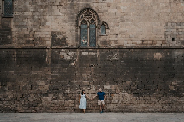 A wide view of the couple standing below a tall historic stone wall with a Gothic window in Barcelona’s Gothic Quarter during their anniversary photoshoot in Barcelona.