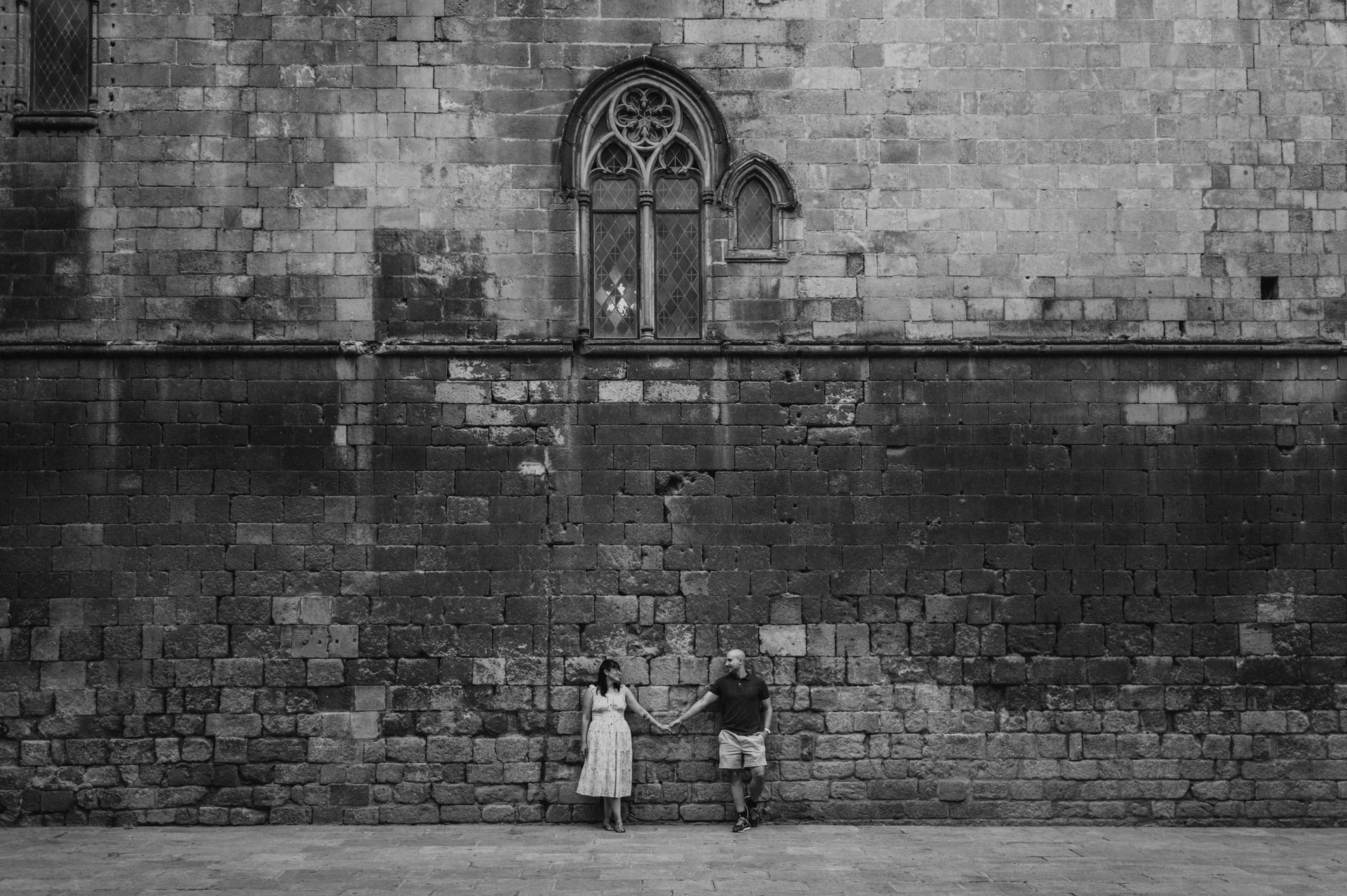 Black-and-white photograph: a wide view of the couple standing below a tall historic stone wall with a Gothic window in Barcelona’s Gothic Quarter during their anniversary photoshoot in Barcelona.