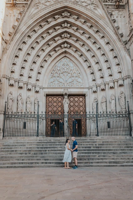 The grand entrance of Barcelona Cathedral around them | Barcelona anniversary photoshoot The couple embracing at the foot of the broad steps beneath Barcelona Cathedral’s ornate entrance during their anniversary photoshoot in Barcelona.