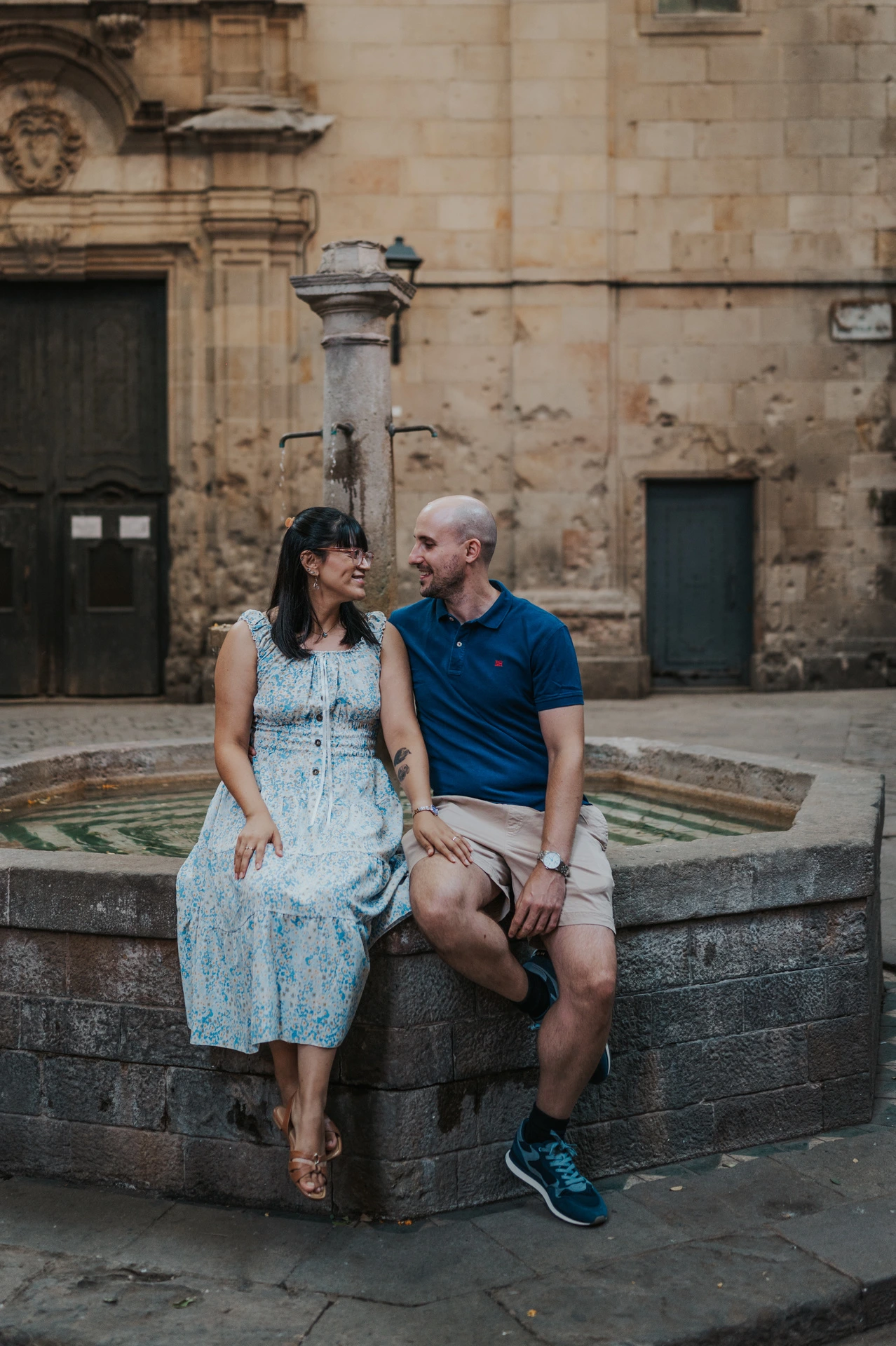 The couple sitting together on the stone edge of the fountain in Plaça de Sant Felip Neri during their anniversary photoshoot in Barcelona.