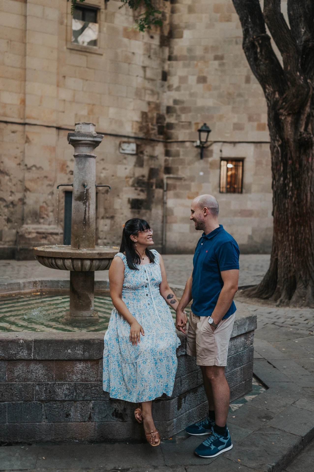 The couple standing hand in hand beside the stone fountain in Plaça de Sant Felip Neri during their anniversary photoshoot in Barcelona.