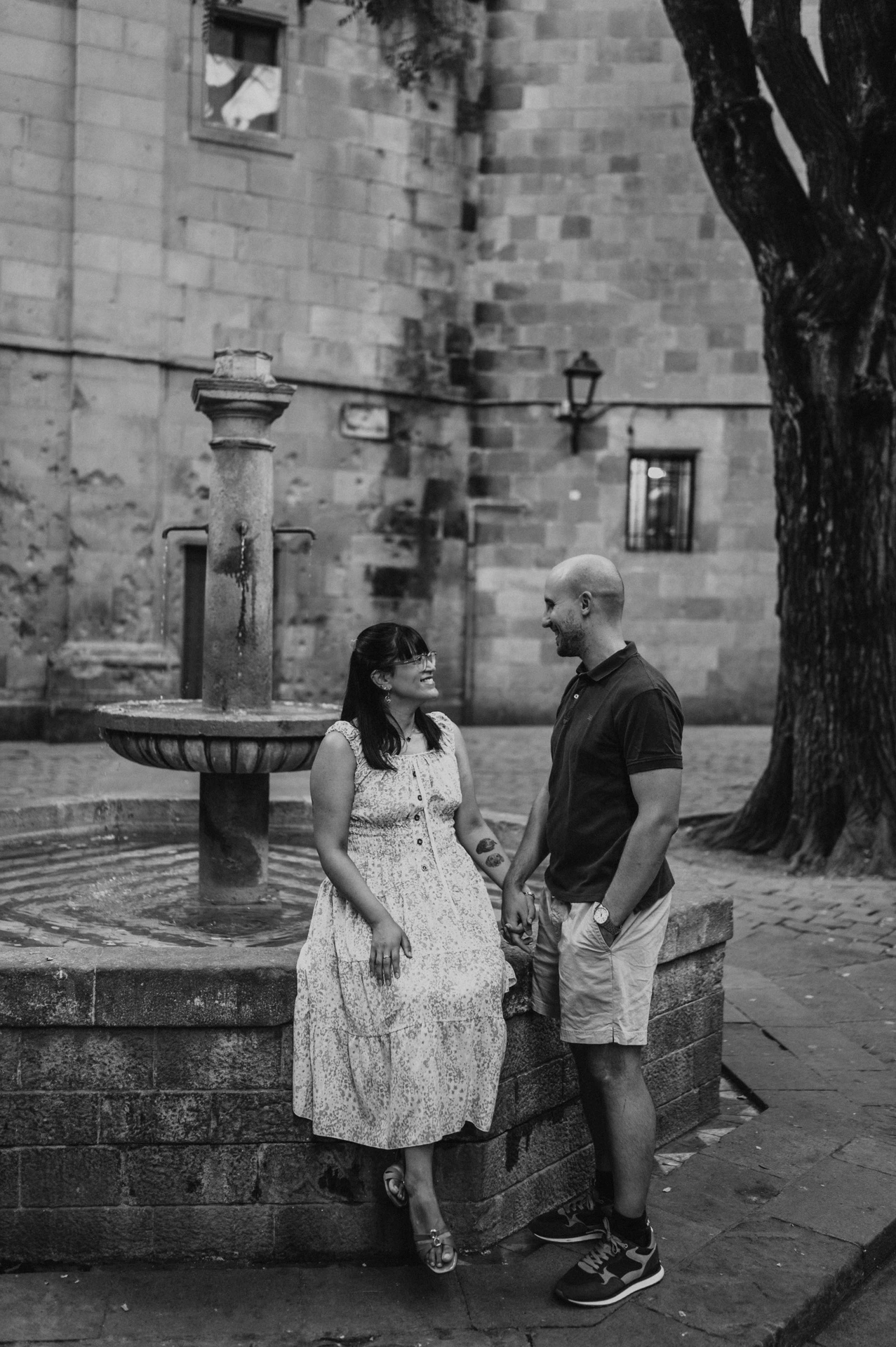 Black-and-white photograph: the couple standing hand in hand beside the stone fountain in Plaça de Sant Felip Neri during their anniversary photoshoot in Barcelona.