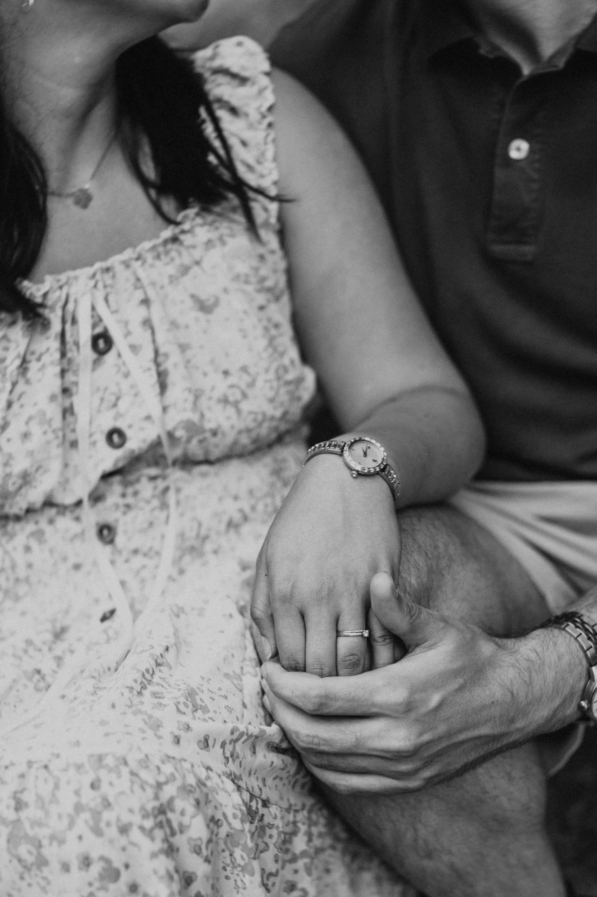 A small hand-in-hand detail from their anniversary session in black and white | Barcelona anniversary photoshoot Black-and-white photograph: a close-up of the couple’s hands resting together, with her floral dress and his polo shirt visible during their anniversary photoshoot in Barcelona.
