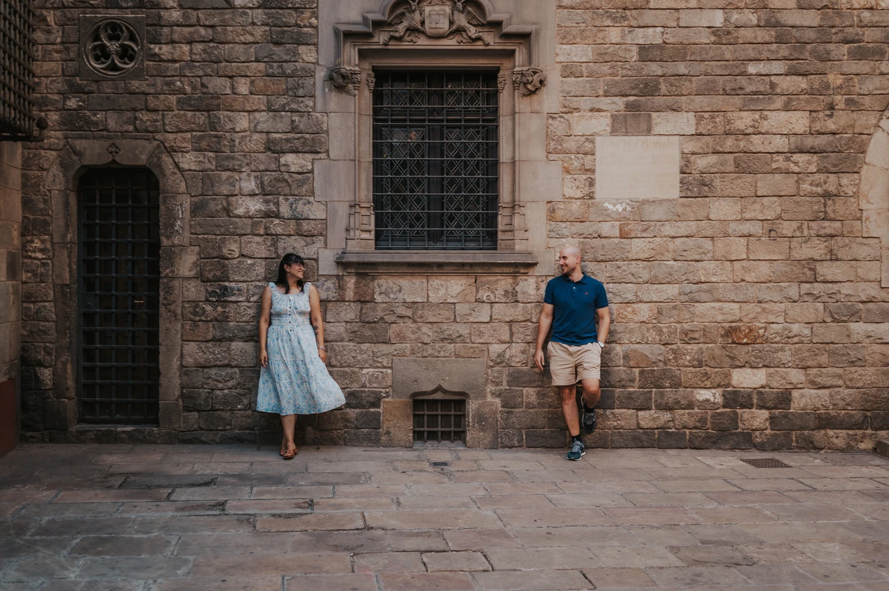 Old stone and a carved window around them | Barcelona anniversary photoshoot The couple standing on either side of a barred historic window in a textured stone wall in the Gothic Quarter during their anniversary photoshoot in Barcelona.