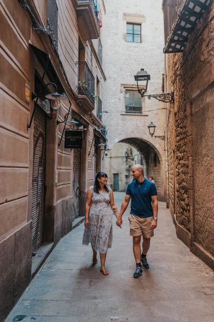 A walk through a narrow old-town lane | Barcelona anniversary photoshoot The couple walking hand in hand through a narrow Gothic Quarter lane with a stone arch behind them during their anniversary photoshoot in Barcelona.