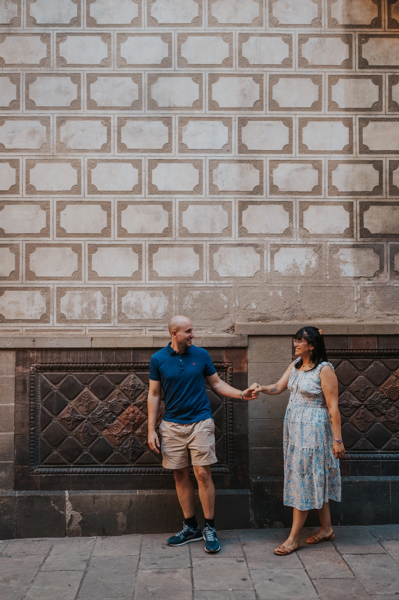 The couple holding hands with space between them beside a patterned stone wall in the Gothic Quarter during their anniversary photoshoot in Barcelona.
