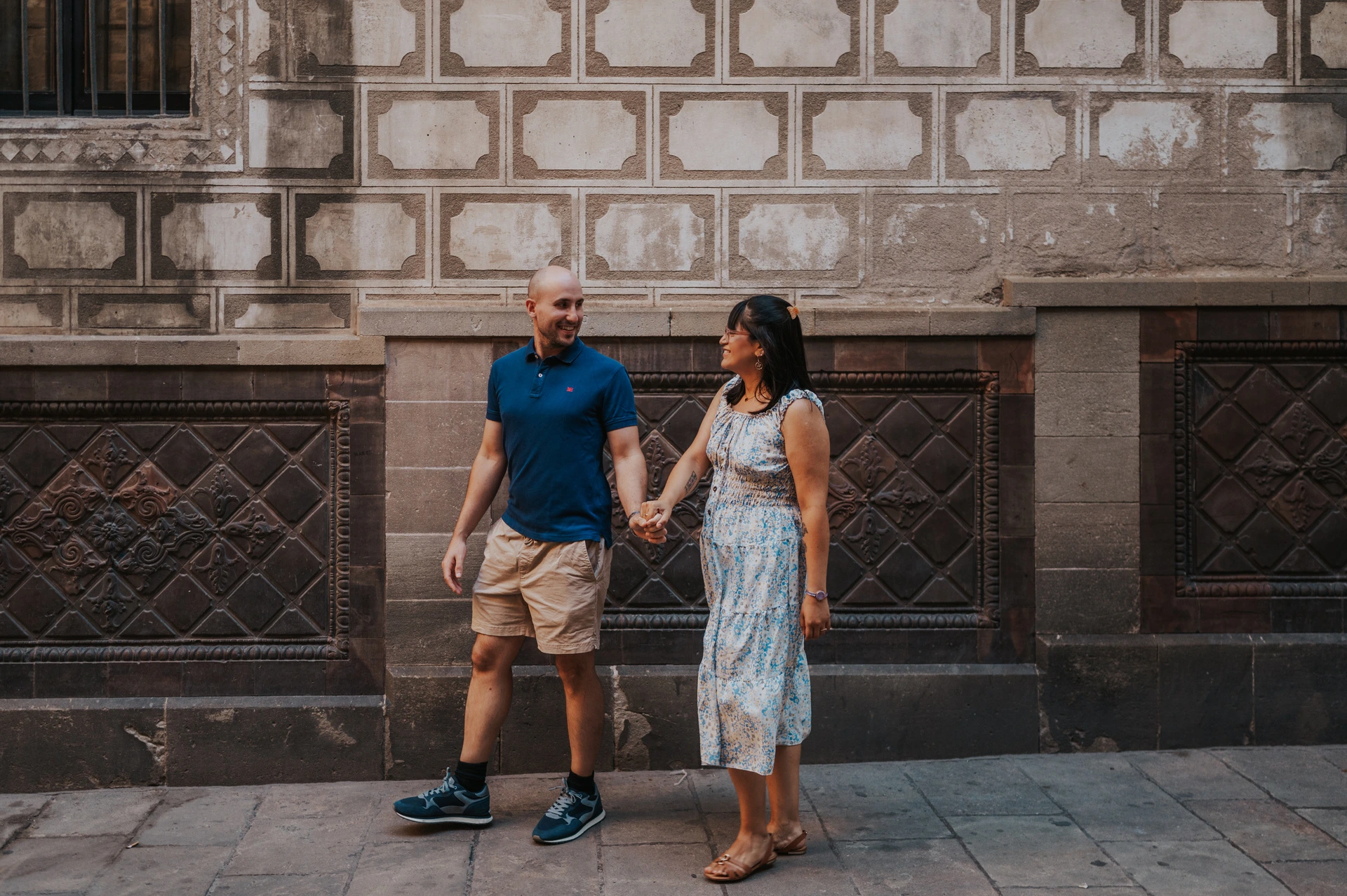 Hand in hand against a patterned old-town wall | Barcelona anniversary photoshoot The couple walking hand in hand beside a patterned stone wall in the Gothic Quarter during their anniversary photoshoot in Barcelona.