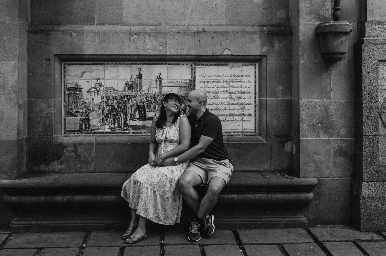 A close moment on a Gothic Quarter stone bench in black and white | Barcelona anniversary photoshoot Black-and-white photograph: the couple sitting close together on a stone bench beneath a decorative picture panel in the Gothic Quarter during their anniversary photoshoot in Barcelona.
