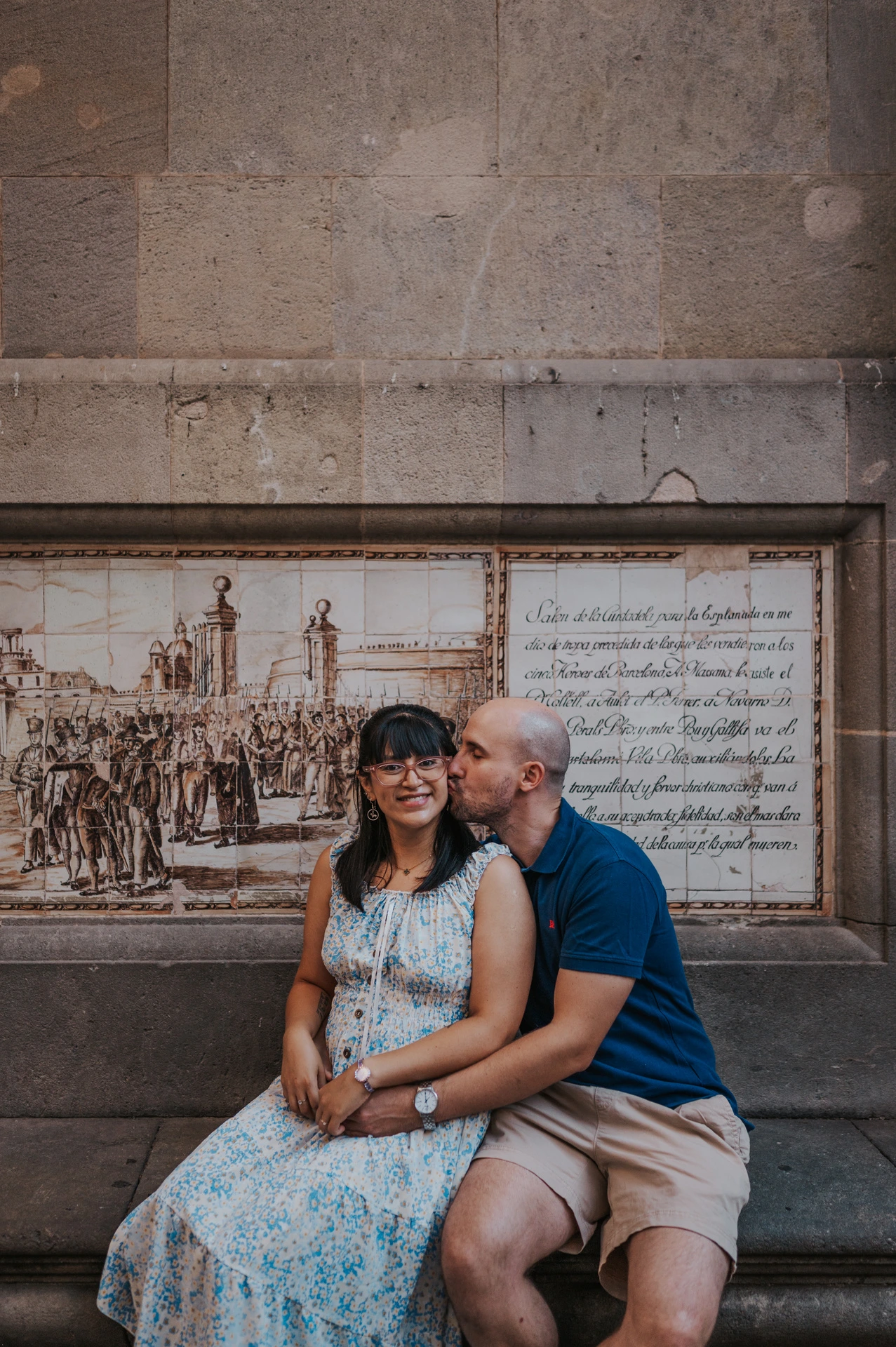 A close portrait of the man kissing his partner’s cheek as they sit on a stone bench beneath a decorative picture panel in the Gothic Quarter during their anniversary photoshoot in Barcelona.