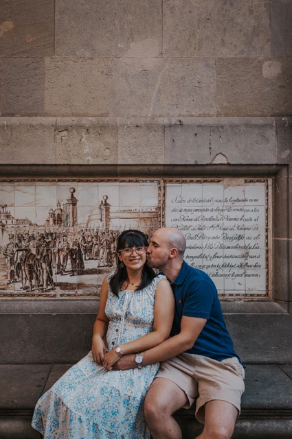 A close moment on a Gothic Quarter stone bench | Barcelona anniversary photoshoot A close portrait of the man kissing his partner’s cheek as they sit on a stone bench beneath a decorative picture panel in the Gothic Quarter during their anniversary photoshoot in Barcelona.