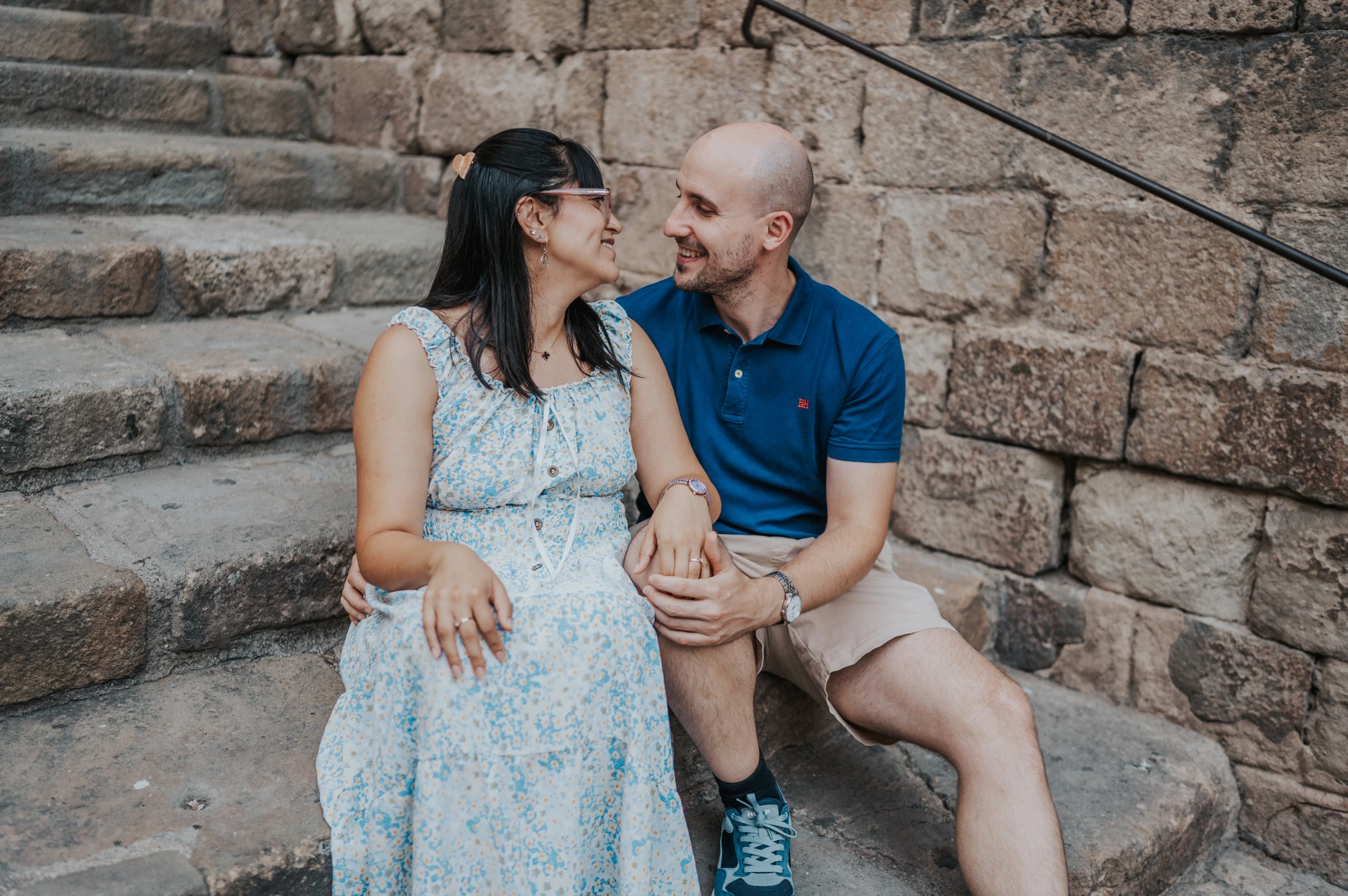 The couple sitting close together on broad stone steps beside a historic wall in the Gothic Quarter during their anniversary photoshoot in Barcelona.