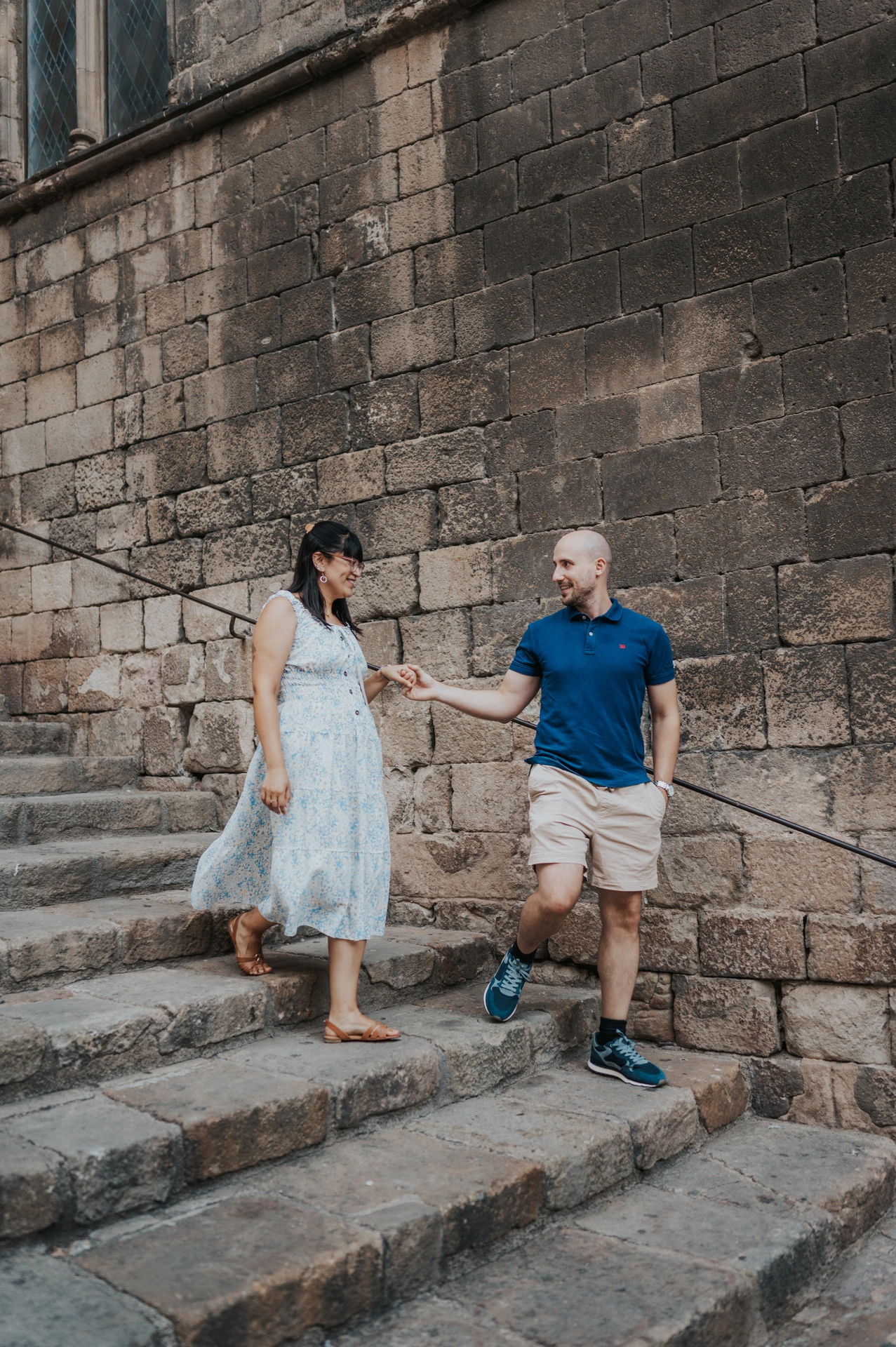 The couple holding hands as they walk down broad stone steps beside a historic wall in the Gothic Quarter during their anniversary photoshoot in Barcelona.