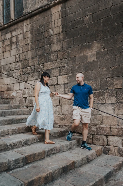 A hand-in-hand walk down the old stone steps | Barcelona anniversary photoshoot The couple holding hands as they walk down broad stone steps beside a historic wall in the Gothic Quarter during their anniversary photoshoot in Barcelona.