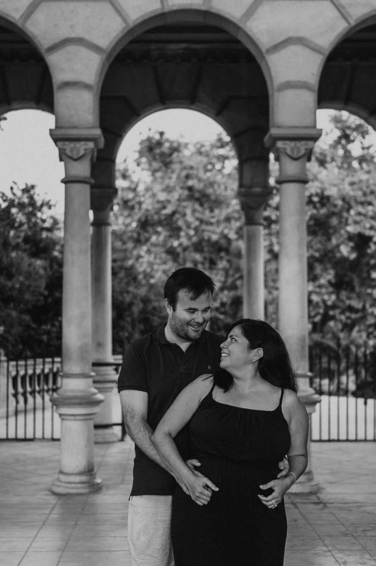 Black-and-white photograph: the parents standing close together beneath the stone arches during their family photoshoot in Barcelona.