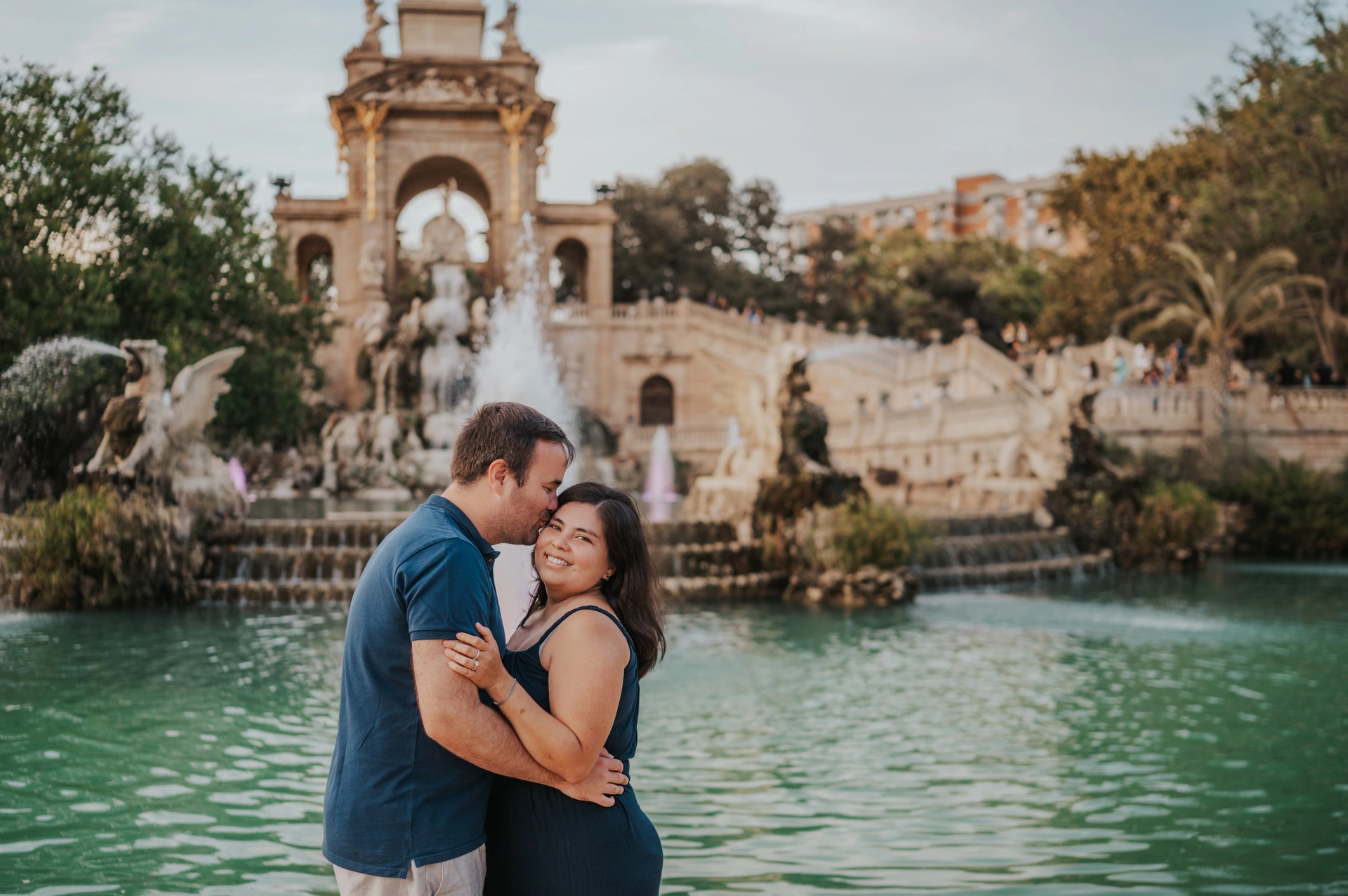 The father leaning towards the mother for a forehead kiss beside the fountain during their family photoshoot in Barcelona.