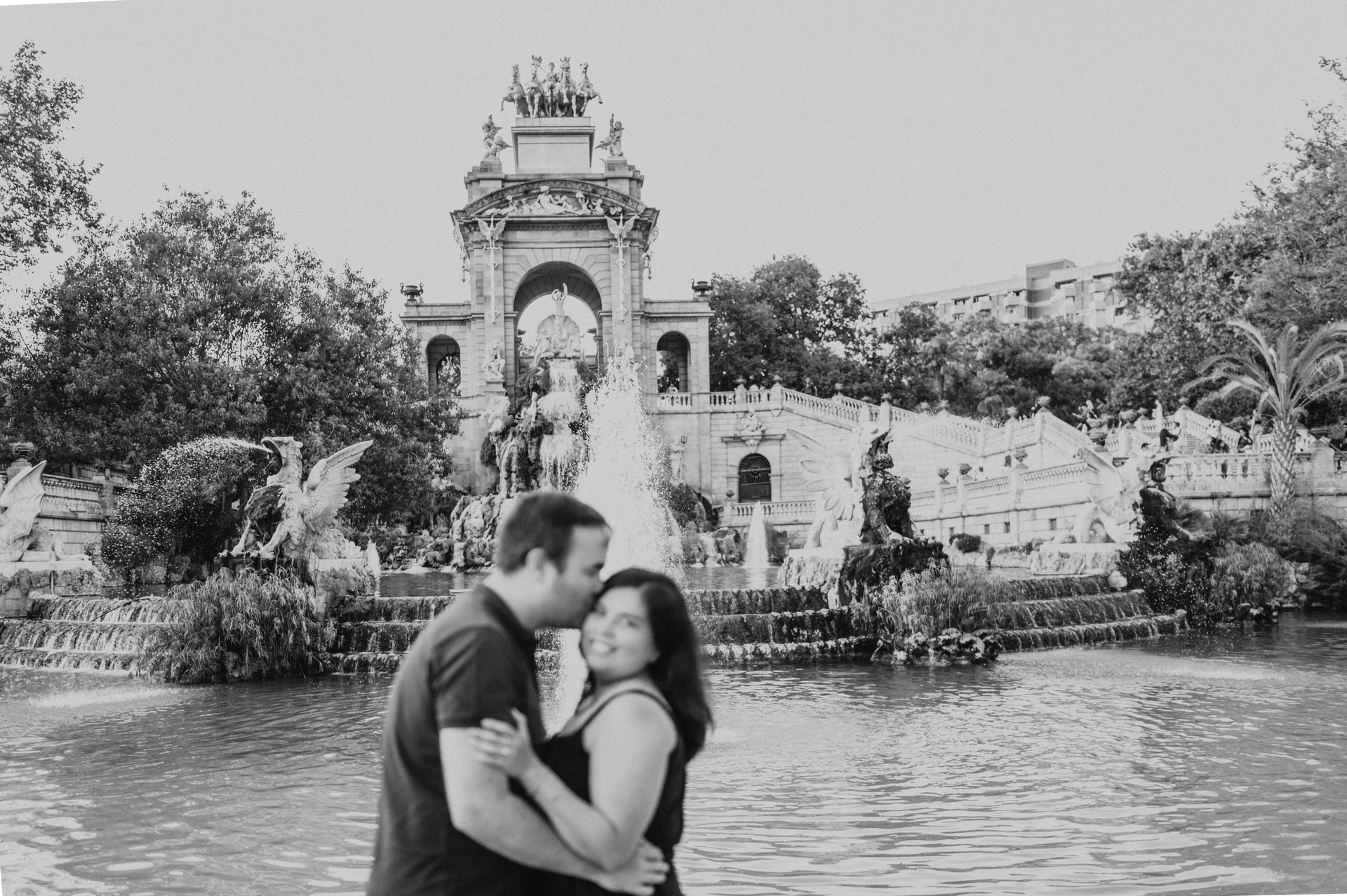 Black-and-white photograph: the father leaning towards the mother for a forehead kiss beside the fountain during their family photoshoot in Barcelona.