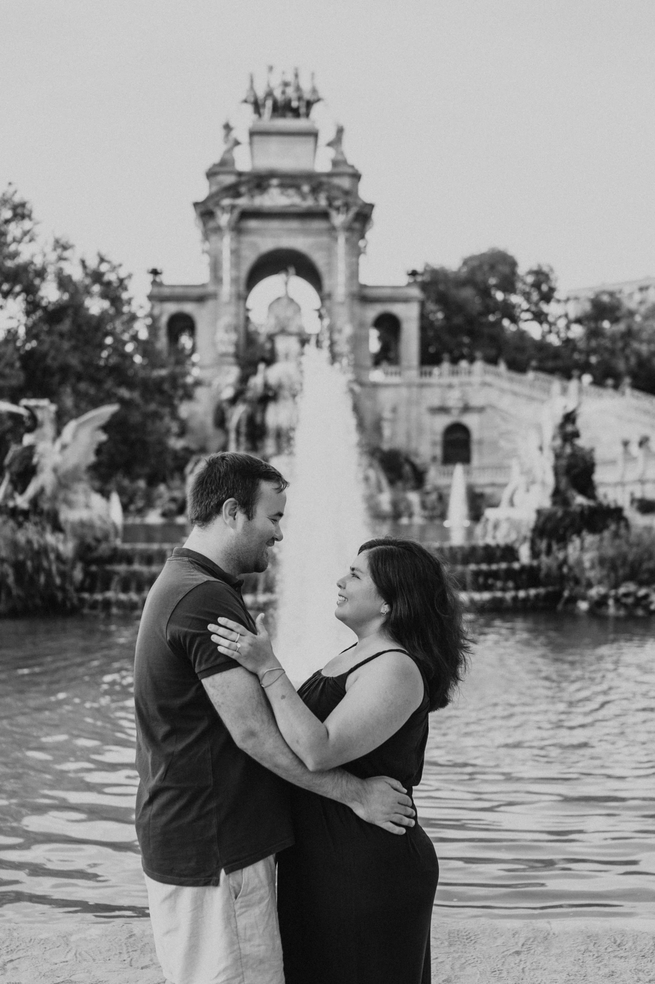 Black-and-white photograph: the parents smiling towards one another beside the fountain during their family photoshoot in Barcelona.