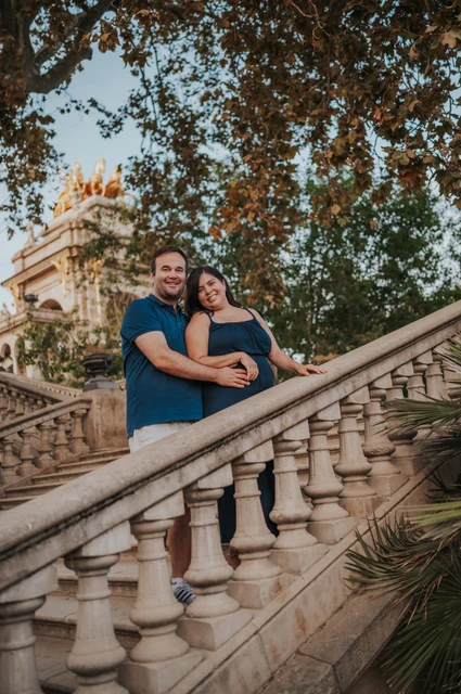 The parents embracing beside the sloping stone balustrade with the fountain behind them during their family photoshoot in Barcelona.