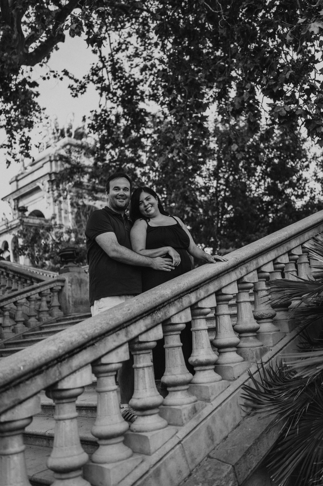 Black-and-white photograph: the parents embracing beside the sloping stone balustrade with the fountain behind them during their family photoshoot in Barcelona.