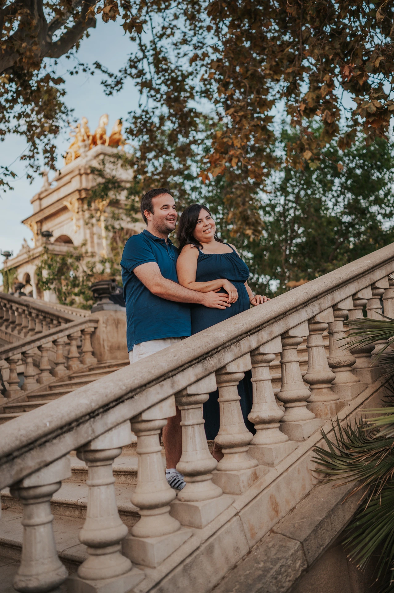 The parents embracing beside the sloping stone balustrade with the fountain behind them during their family photoshoot in Barcelona.
