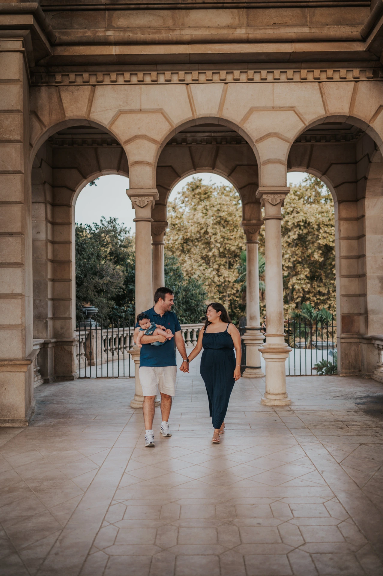 The parents walking together beneath the stone arches while the father carries their child during their family photoshoot in Barcelona.