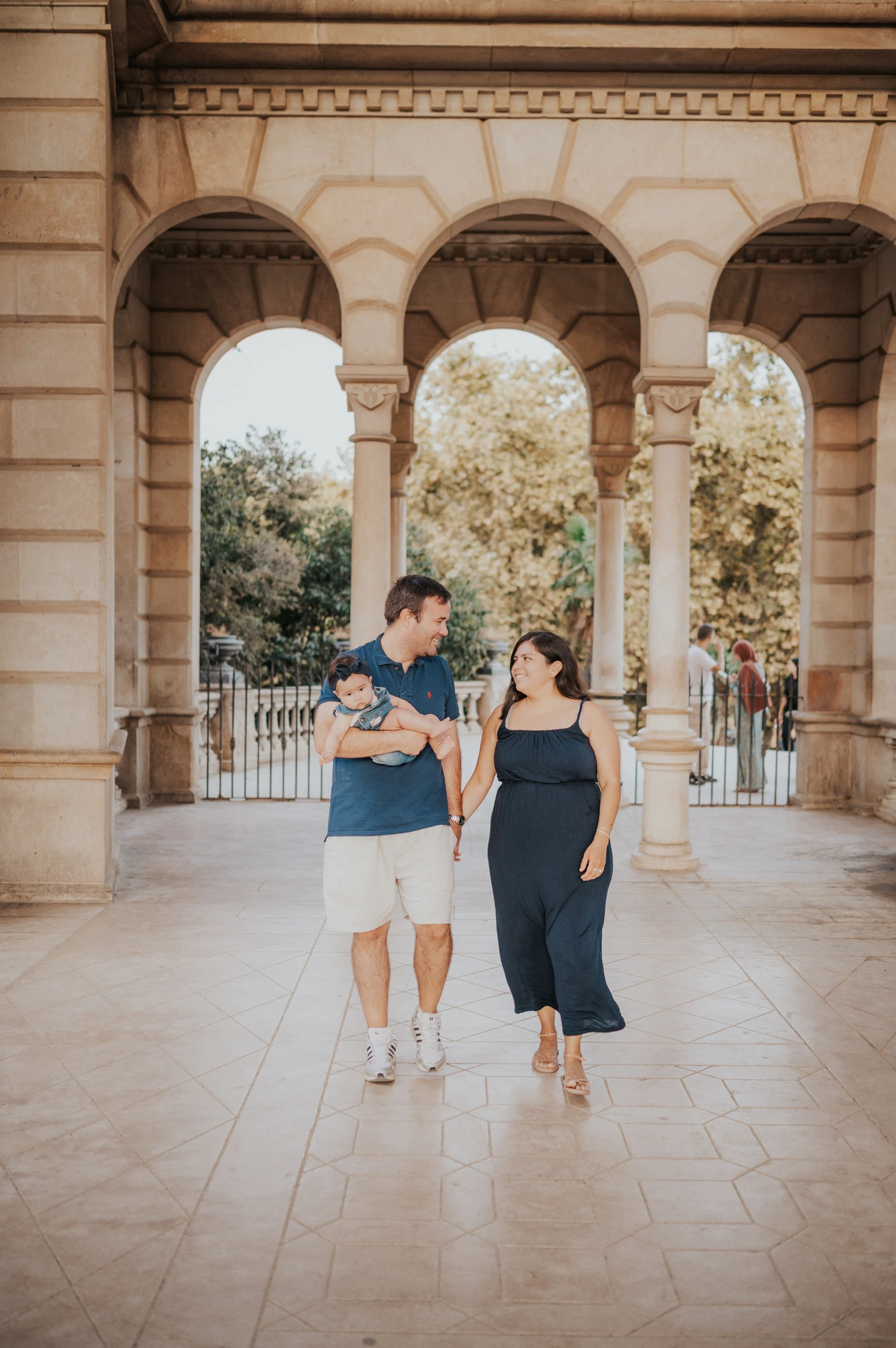 The parents walking together beneath the stone arches while the father carries their child during their family photoshoot in Barcelona.