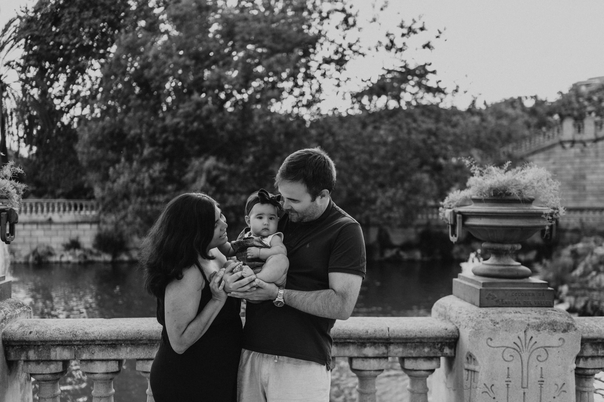 Black-and-white photograph: the parents leaning towards their child beside the stone balustrade during their family photoshoot in Barcelona.