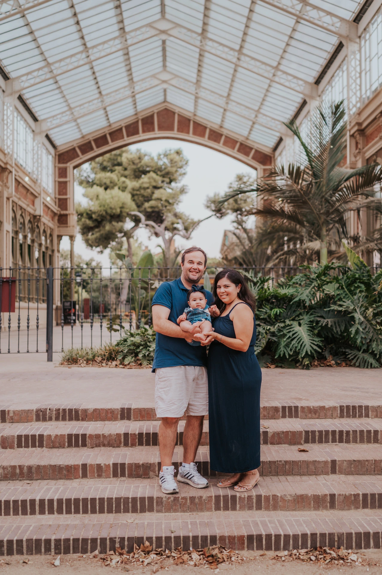The family standing together beneath the glass and iron roof among green plants during their family photoshoot in Barcelona.
