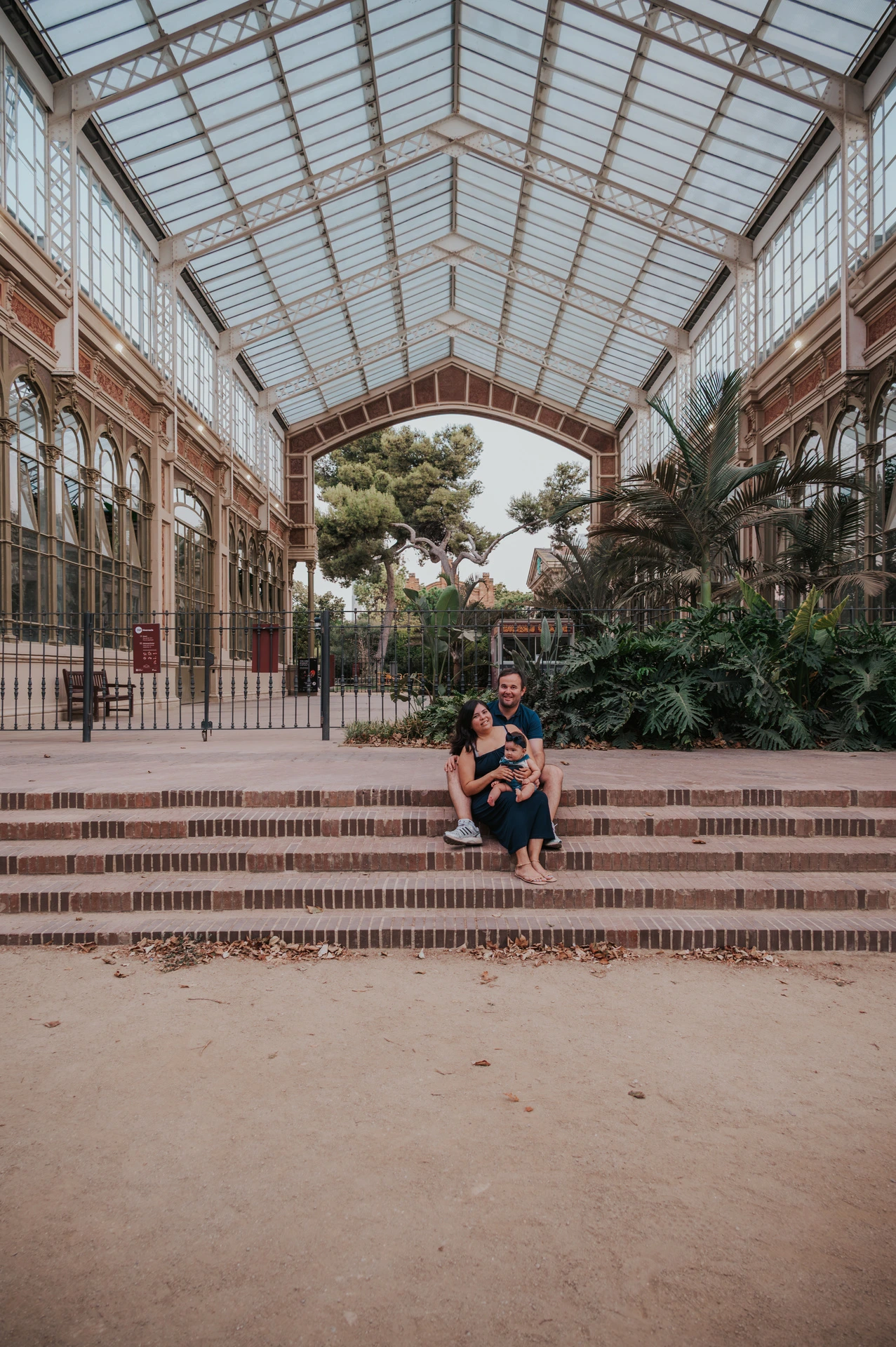 The family standing together beneath the glass and iron roof among green plants during their family photoshoot in Barcelona.