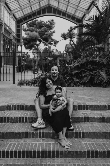 A family portrait on the glasshouse steps in black and white | Barcelona family photoshoot Black-and-white photograph: the family sitting together on the steps beneath a glass and iron roof during their family photoshoot in Barcelona.
