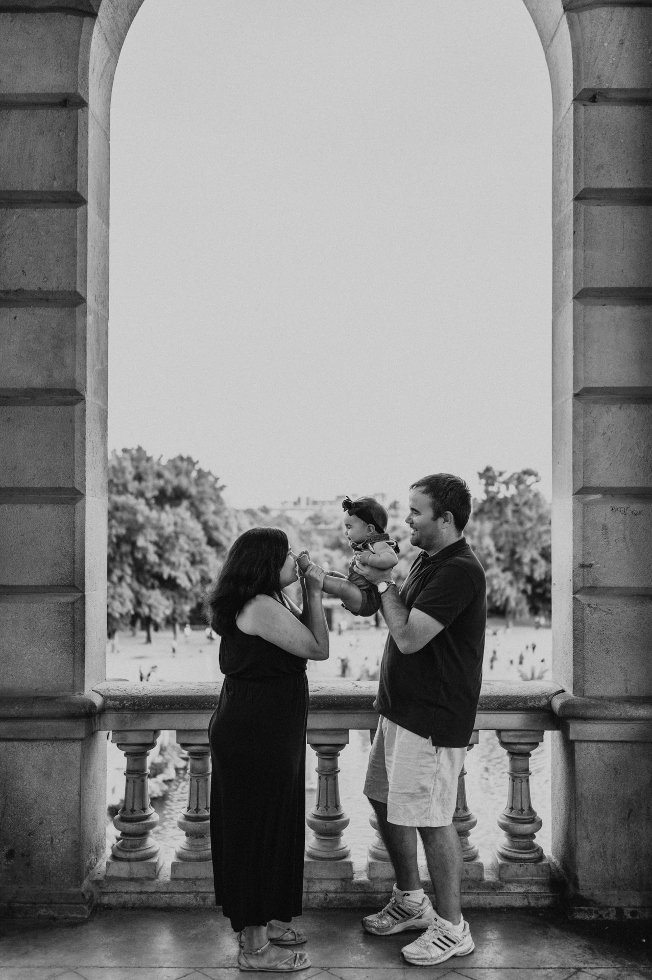 Black-and-white photograph: the parents raising their child between them beneath a stone arch during their family photoshoot in Barcelona.