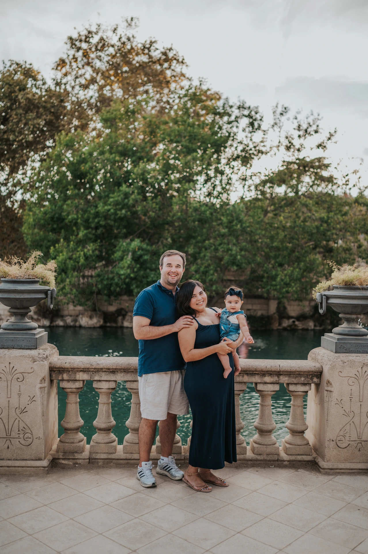 The family standing beside a stone balustrade with trees and water behind them during their family photoshoot in Barcelona.