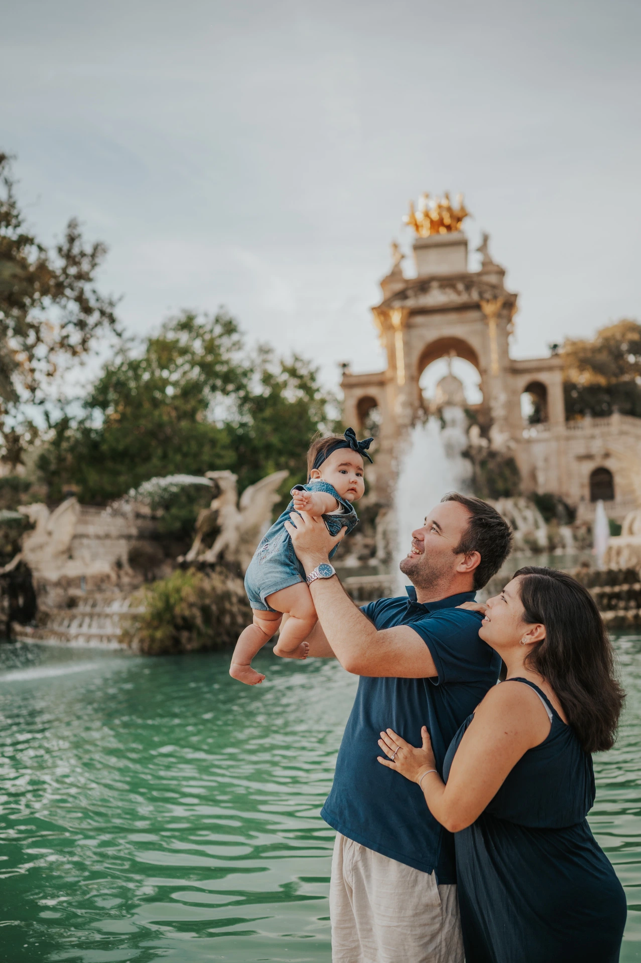 The father lifting his child while the mother looks up at them beside the fountain during their family photoshoot in Barcelona.