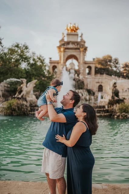 The father lifting his child while the mother looks up at them beside the fountain during their family photoshoot in Barcelona.