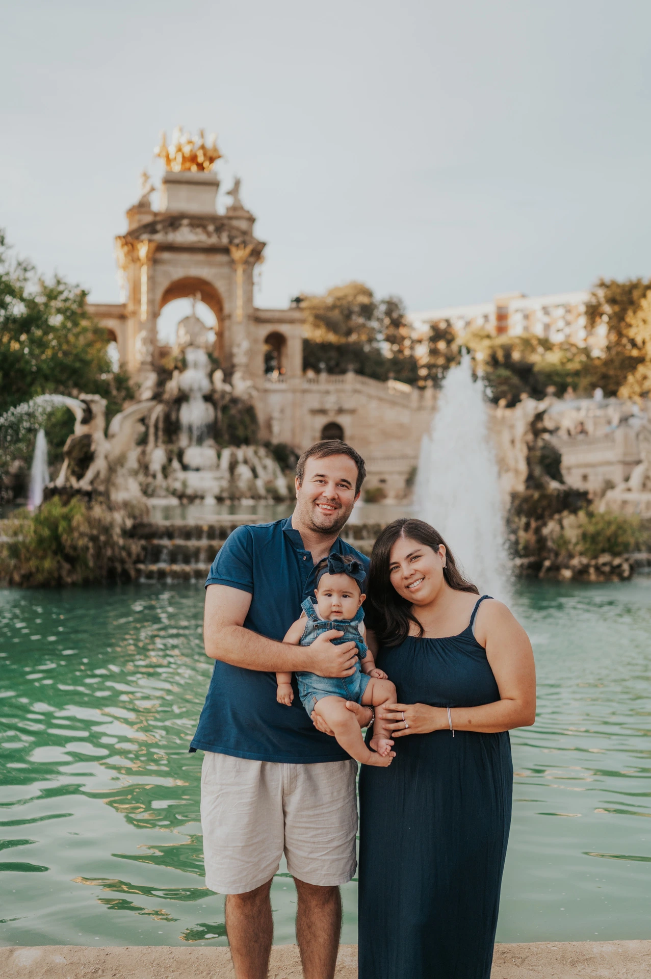 The parents holding their one-year-old together in front of the fountain and green water during their family photoshoot in Barcelona.