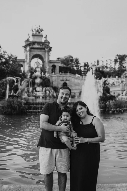A family portrait beside the fountain in black and white | Barcelona family photoshoot Black-and-white photograph: the parents holding their one-year-old together in front of the fountain and green water during their family photoshoot in Barcelona.