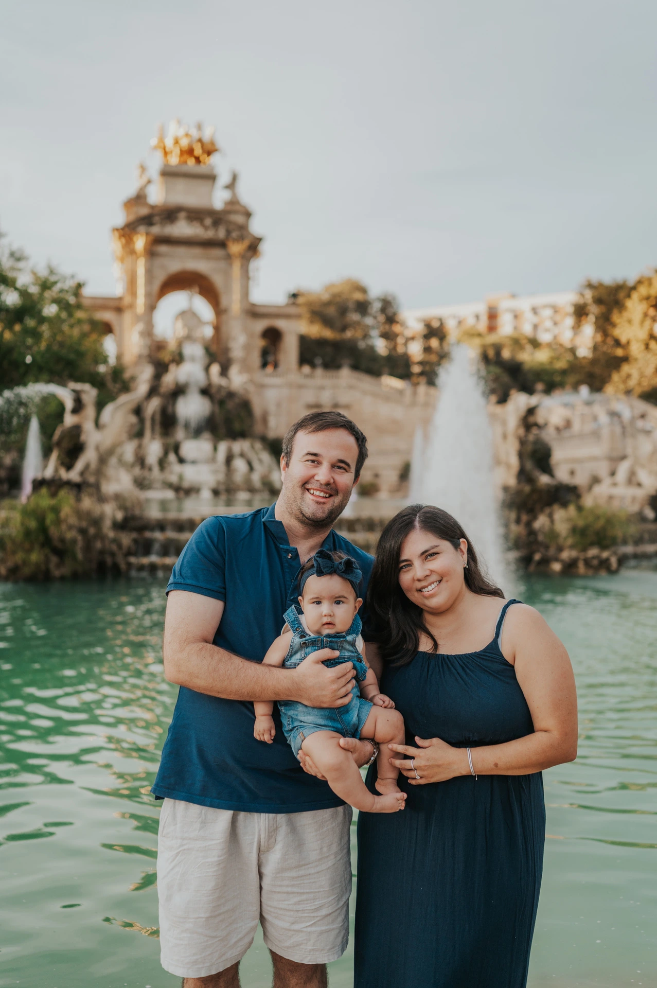The parents holding their one-year-old together in front of the fountain and green water during their family photoshoot in Barcelona.