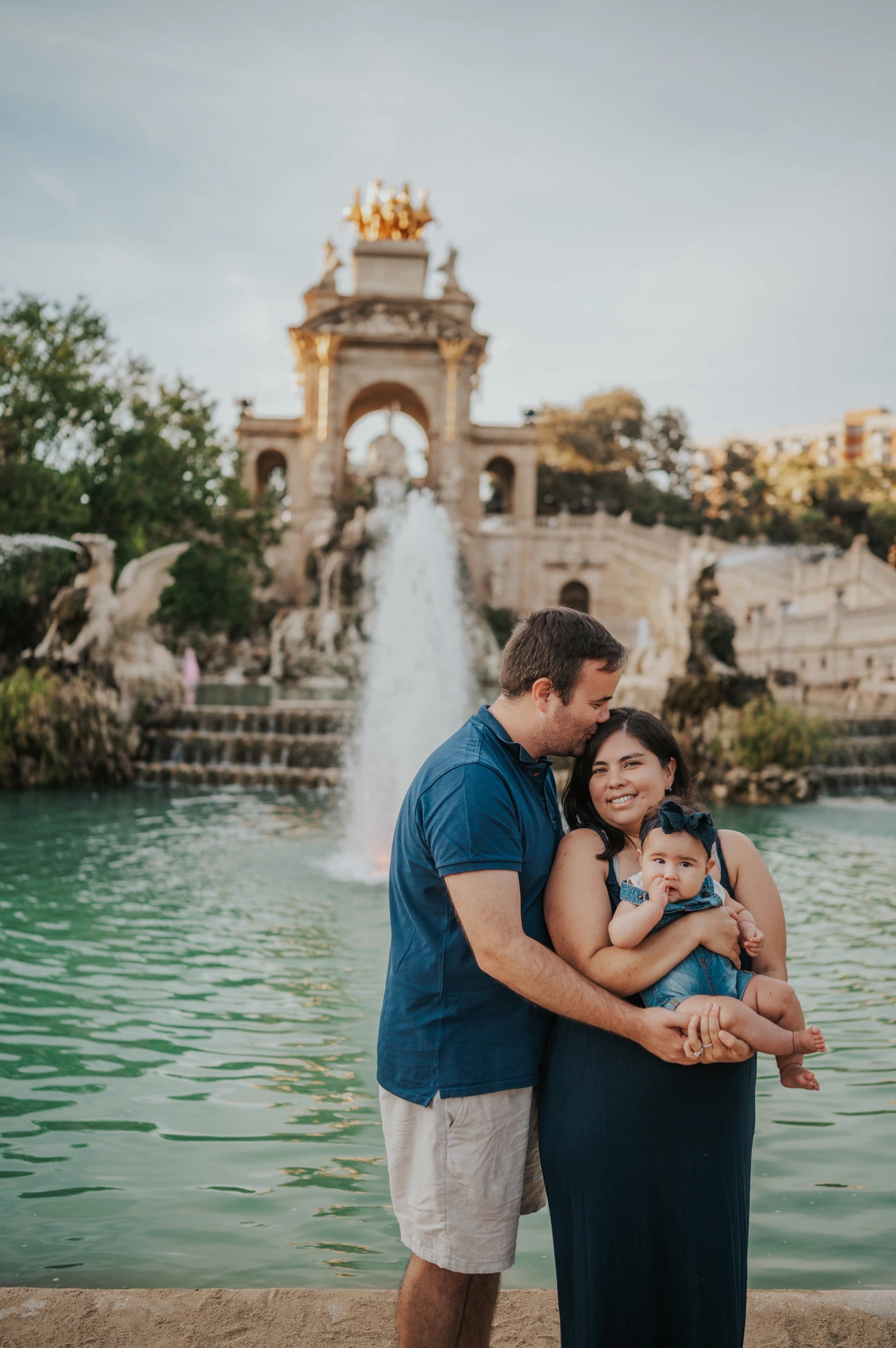 The mother holding their child while the father leans in close beside the fountain during their family photoshoot in Barcelona.
