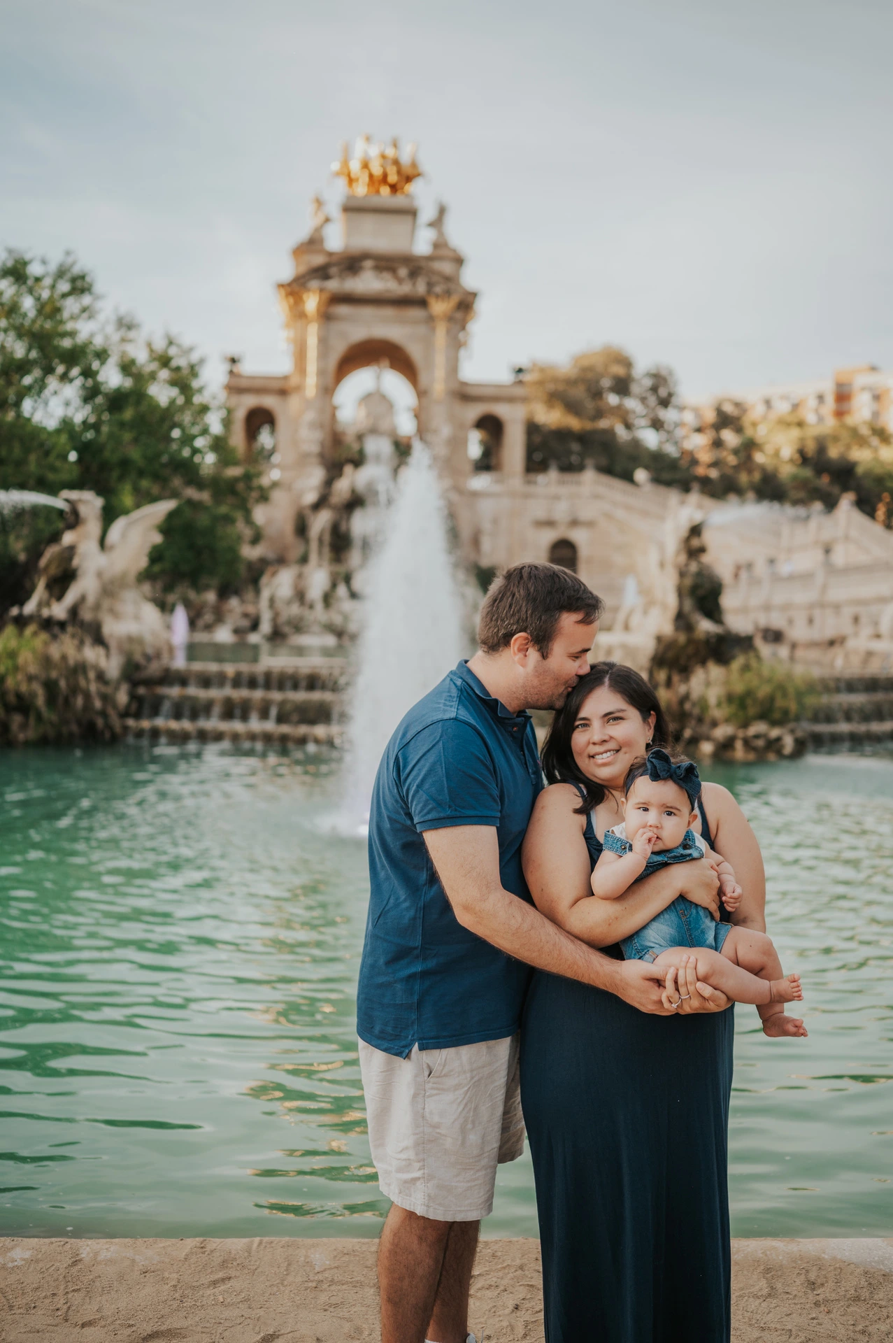 The mother holding their child while the father leans in close beside the fountain during their family photoshoot in Barcelona.