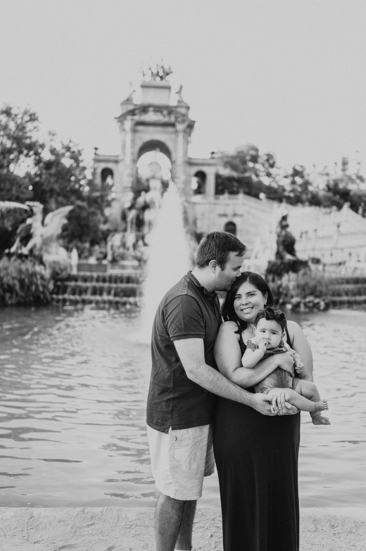 Black-and-white photograph: the mother holding their child while the father leans in close beside the fountain during their family photoshoot in Barcelona.