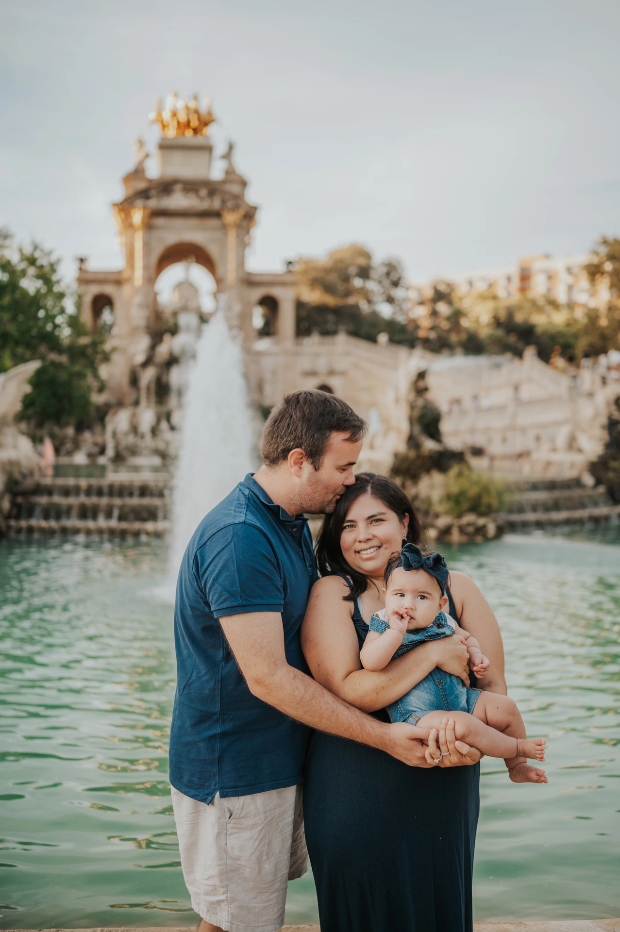The mother holding their child while the father leans in close beside the fountain during their family photoshoot in Barcelona.