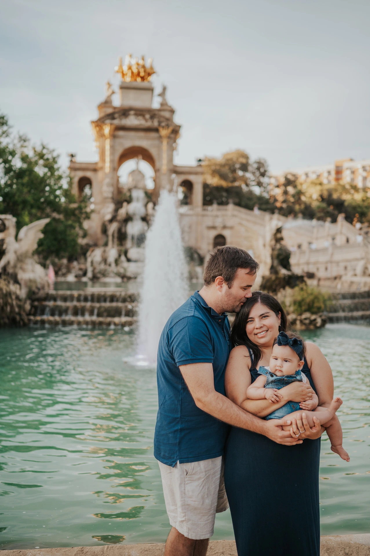 The mother holding their child while the father leans in close beside the fountain during their family photoshoot in Barcelona.