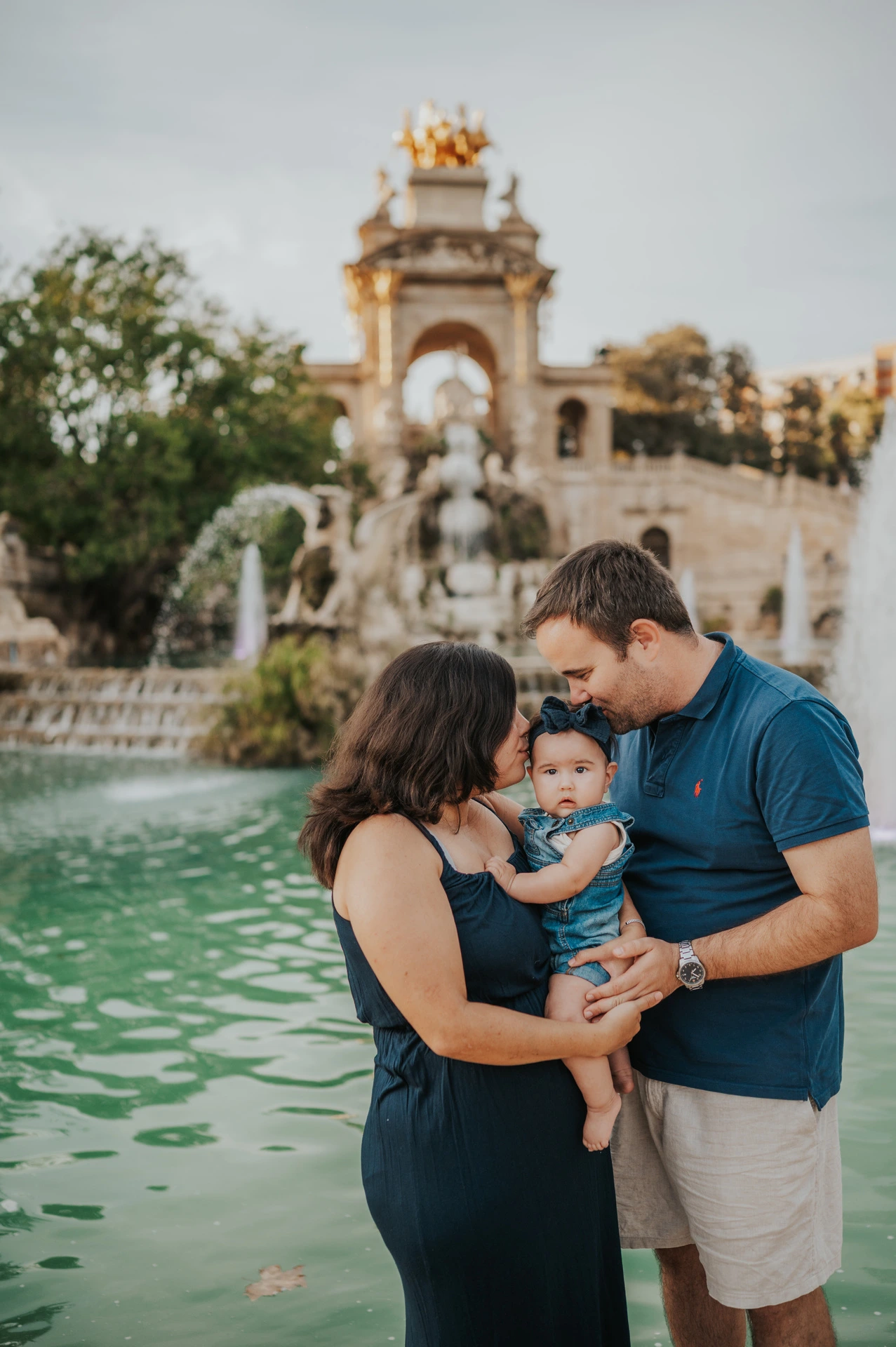 Both parents leaning close to kiss their child beside the water during their family photoshoot in Barcelona.