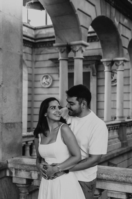 Black-and-white photograph: the couple embracing beneath a stone arch beside the carved railing during their engagement photoshoot in Barcelona.